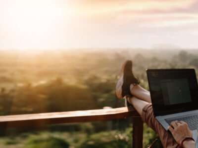 A host sits on a wooden deck at sunset, laptop in lap and feet up on the railing, looking out across tree-lined hills.