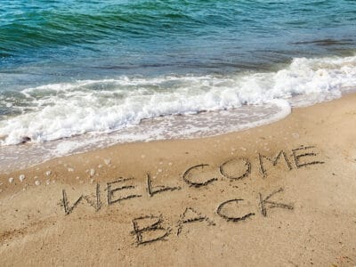 The words 'Welcome back' drawn in beach sand beside the sea, evoking a returning guest's next stay